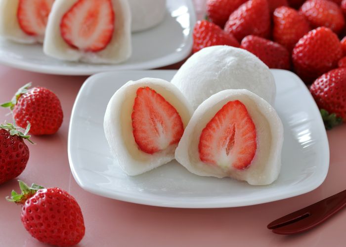 Strawberry daifuku cut in half on a white plate, showing a filling of white bean paste and fresh strawberry surrounded by white mochi.